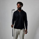 Man holding a golf club wearing a black long-sleeve shirt and beige pants on a gray background