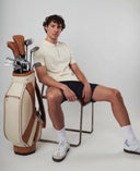 Man sitting on a chair next to a golf bag with golf clubs on a plain background