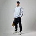 Man wearing a light gray sweatshirt, black pants, white sneakers, and a dark cap, holding a green bag on a plain background.