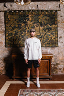 Man standing in a room with stone walls and a large tapestry, wearing a white long-sleeve shirt and black shorts.