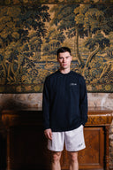 Man wearing a black sweatshirt and white shorts standing in front of an ornate wall tapestry.