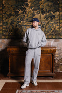 Person wearing a gray hoodie and pants standing in front of an ornate tapestry.